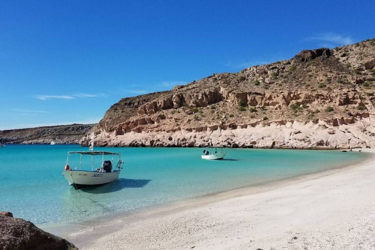 Turquoise Mediterranean beach with anchored fishing boats, white sandy shore, and rocky cliff hillside under clear blue sky