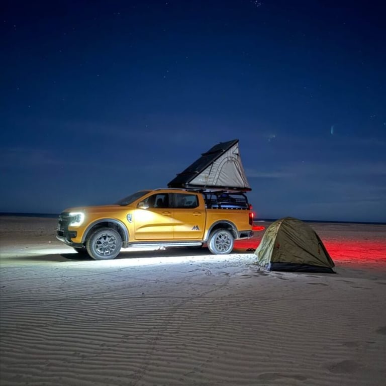 Gold pickup truck with roof tent set up on a beach at night, illuminated by vehicle lights and a tent with red glow