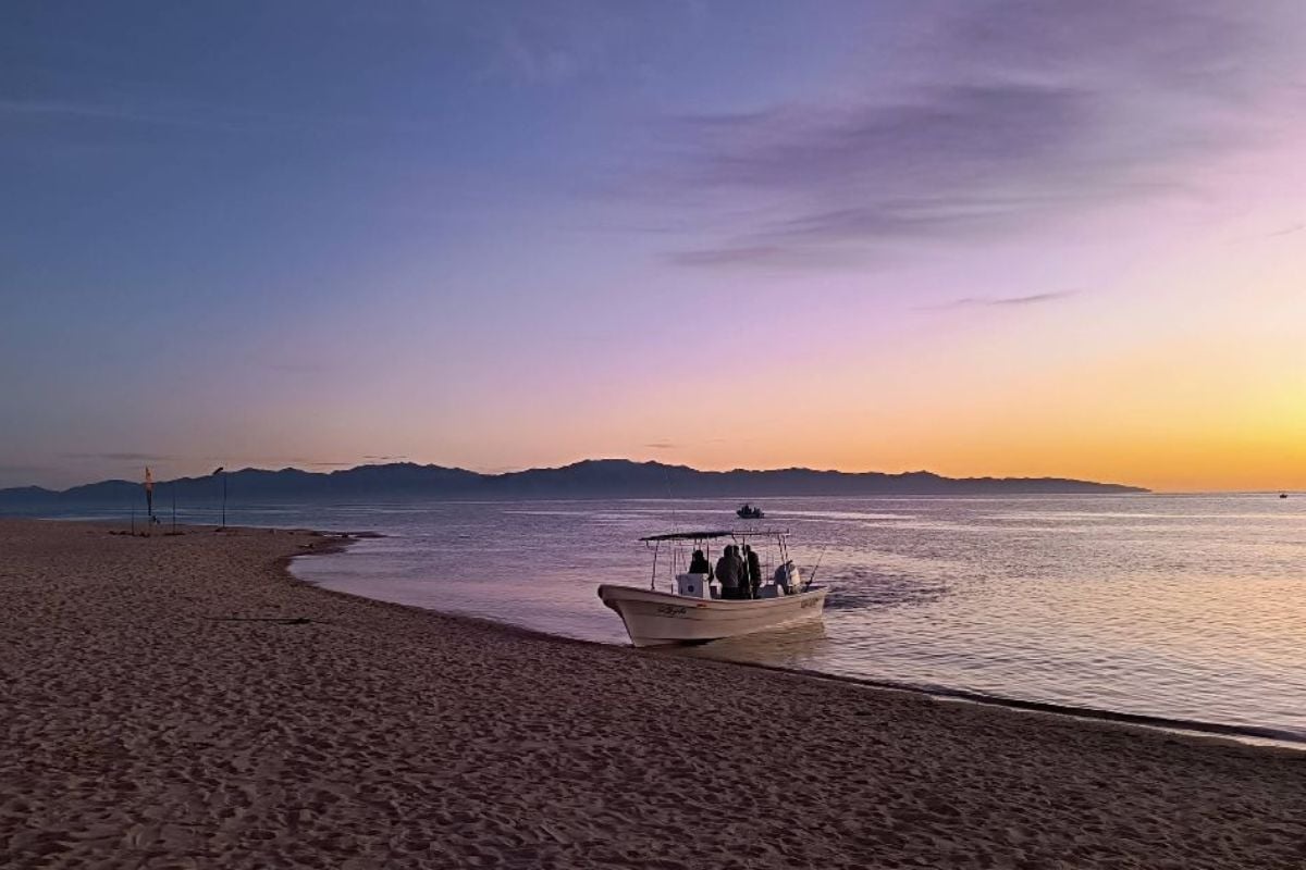 Fishing boat beached at sunset on sandy shore with purple and orange sky over distant mountains