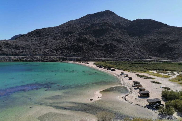 Coastal beach with turquoise water, sandy shore, parked vehicles, and a dark volcanic mountain in the background