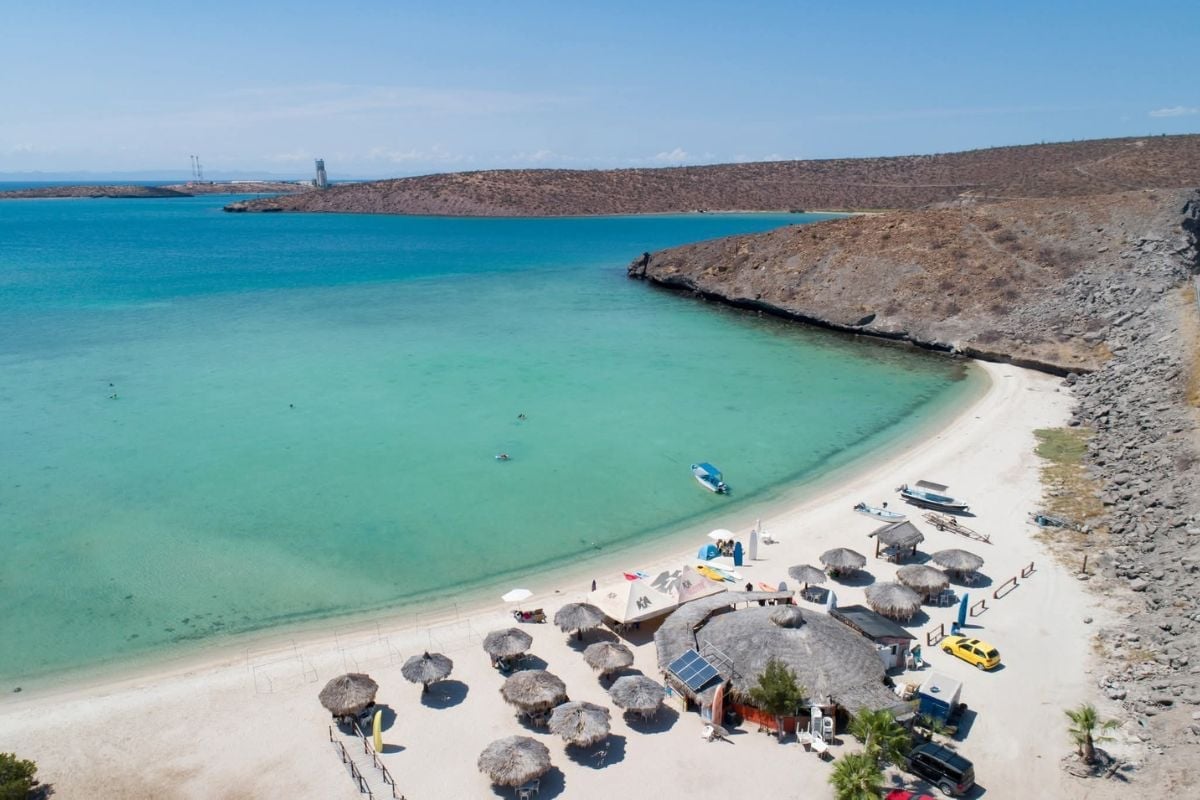 Aerial view of a secluded beach cove with turquoise waters, white sand, and rocky cliffs with beach umbrellas and boats