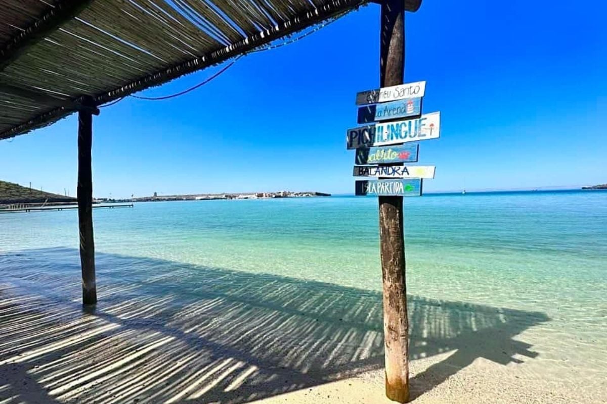Wooden directional signs on a post at a tropical beach with clear turquoise water and wooden dock overhead