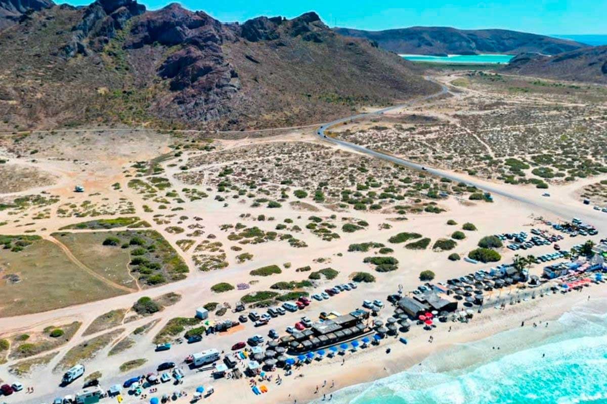 Aerial view of a crowded beach with rows of parked cars, desert landscape, mountains, and turquoise ocean water