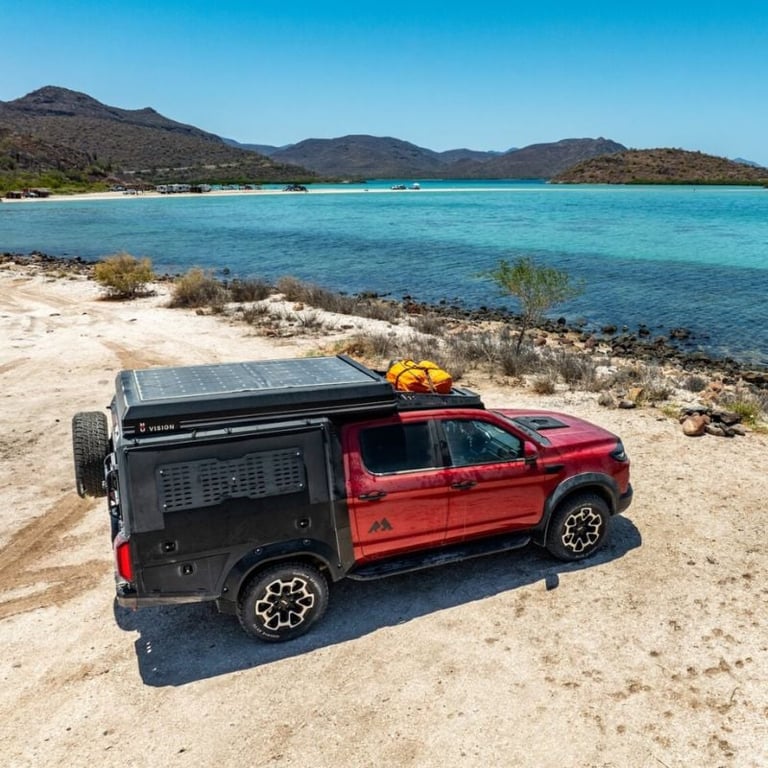 Red pickup truck with roof rack and solar panel parked on sandy beach with turquoise water and mountains in background