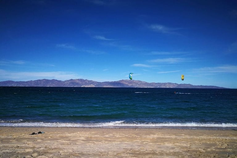 Beach scene with turquoise ocean, sandy shore, distant mountains, and kitesurfers on a clear blue sky day