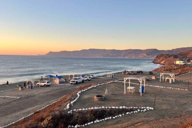 Coastal beach scene at sunset with parked vehicles, decorative white stones on sand, arbor structure, and mountains in the distance