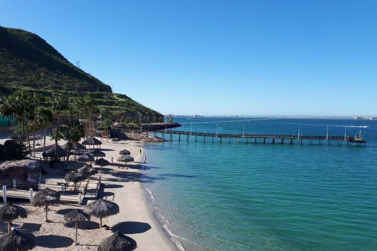 Coastal beach scene with sun umbrellas, sandy shore, turquoise water, wooden pier, and hillside vegetation under clear blue sky
