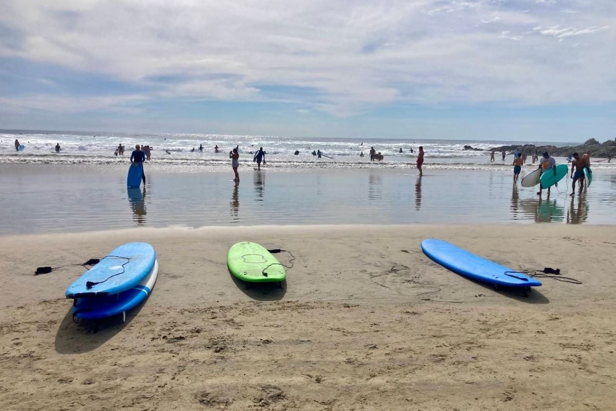 Three surfboards on sandy beach with swimmers and beachgoers in the ocean under a partly cloudy sky