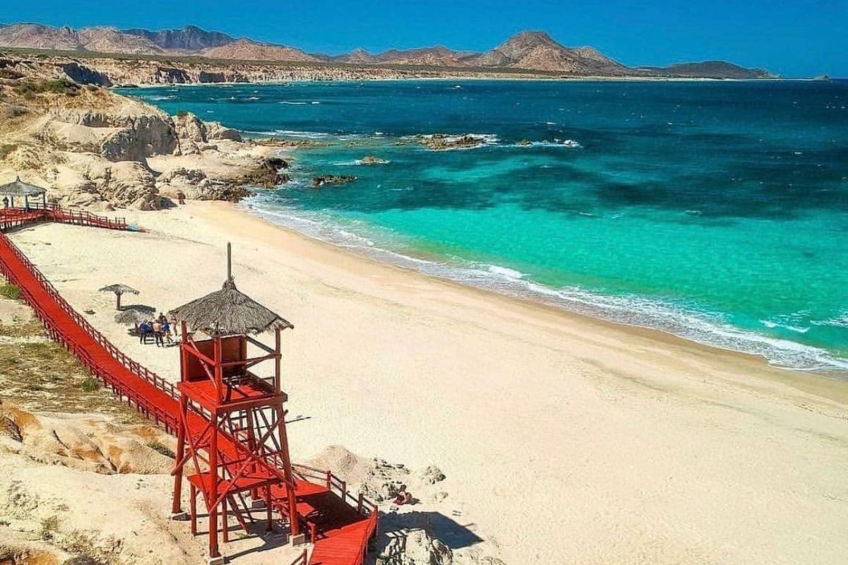 Red lifeguard tower on sandy beach with turquoise water and rocky cliffs under clear blue sky