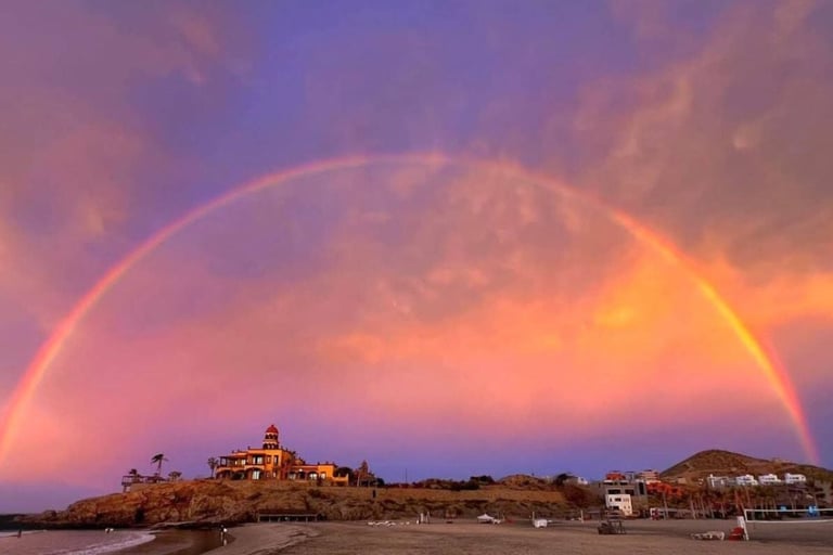 Double rainbow arching over a coastal hillside town with buildings and palm trees at sunset.