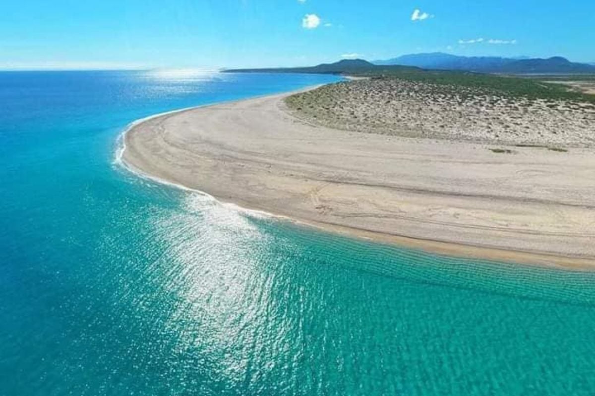 Aerial view of a sandy curved beach with turquoise waters and forested mountains in the distance under blue sky.