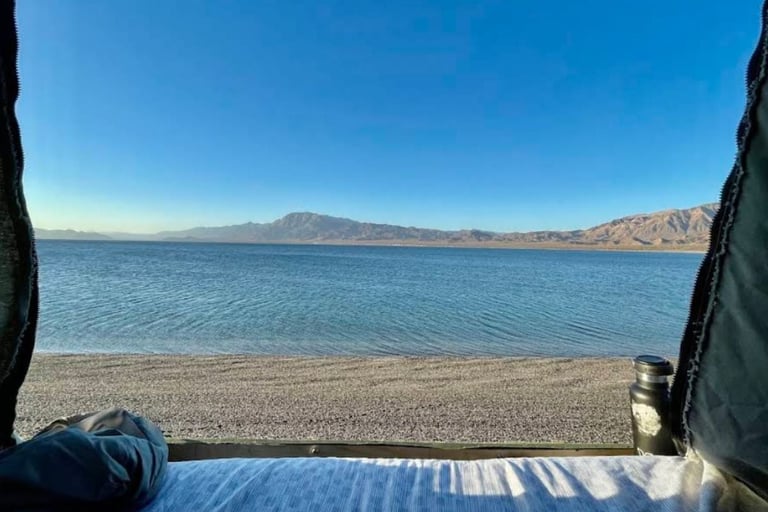 View from inside a tent looking out at a calm turquoise lake with a mountain in the distance and sandy beach
