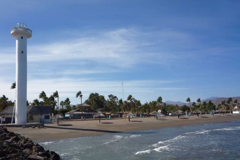 White lighthouse tower on a sandy beach with palm trees, buildings, and ocean waves under a blue sky
